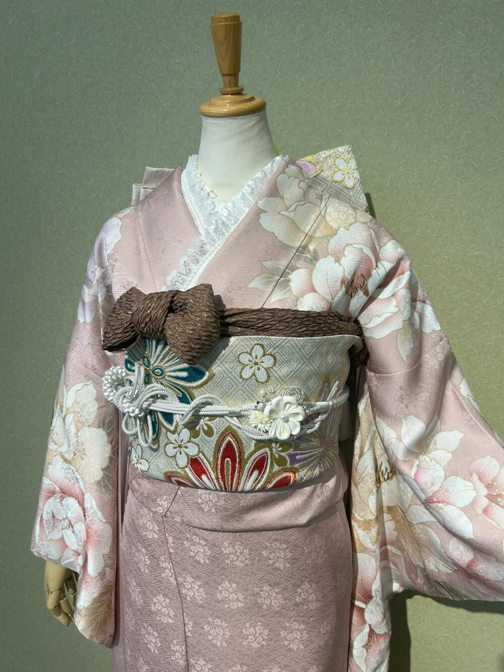 くすみピンクの振袖に、白地に赤や青の古典柄が織り込まれた袋帯、深みのある茶色の帯揚げ、お花の飾りが添えられた白系の帯締め、白のパールとレースがついた重ね衿、淡いゴールド系の半衿を使用したコーディネート