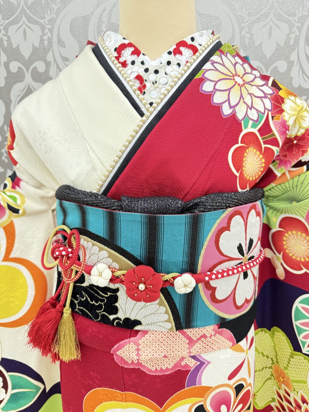 白と赤を基調とした振袖に、鮮やかなターコイズブルーに引き締め効果のある黒の縦縞が大胆に配されている袋帯と、中央に可愛らしい花モチーフの飾りがあしらわれた鮮やかな赤色の帯締めと、シックな黒地に銀通しの帯揚げを使用した振袖コーディネート
